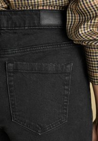 Čierne rifľové nohavice s zadným vreckom so šitým akcentom, textúrovanou tkaninou a koženou visačkou na páse viditeľnou v hornej časti.