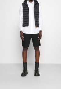 Chaleco acolchado negro con capucha, camisa blanca de botones, pantalones cortos negros y botas negras hasta el tobillo sobre un fondo neutro.