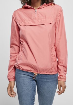 Frau trägt eine pinke Windjacke mit Halbreißverschluss und Kordelzügen sowie blaue Jeans, beschnitten oberhalb des Halses und in der Mitte des Oberschenkels.