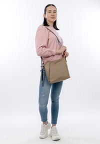 Noor naine roosa pusaga, rebenenud siniste teksade ja valgete tossudega, kandmas pruunikat ristkotti, seismas valge tausta ees.