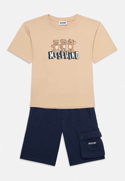 Μπεζ κοντομάνικη μπλούζα με τρεις αρκουδάκια πάνω από το κείμενο «MOSCHINO», συνδυασμένη με σκούρο μπλε σορτσάκι που διαθέτει τσέπη με το λογότυπο «MOSCHINO».