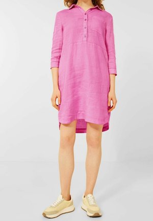 Kobieta w luźnej lnianej sukience w kolorze różowym z guzikami, z rękawami do łokci, ubrana w beżowe trampki, stoi na tle jednolitego białego tła.