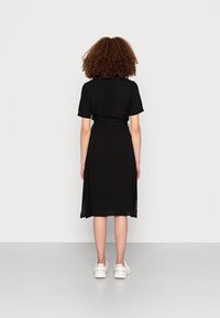 Un vestito nero con maniche corte, vita strizzata, gonna di lunghezza al ginocchio e un sovrapposizione trasparente. Indossato con sneakers bianche, mostrando una texture liscia.