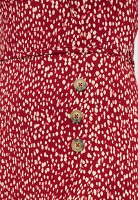 Κόκκινο ύφασμα με ακανόνιστο μοτίβο λευκών κουκκίδων και τρία κάθετα ευθυγραμμισμένα μπεζ κουμπιά με κόκκινες ραφές κλωστής.