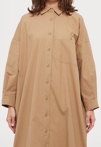 Vestido camisetón marrón de botones, fabricado en algodón, que presenta un bolsillo, un cuello y mangas largas con un corte holgado. Textura suave, diseño sencillo.