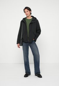 Mężczyzna w czarnej kurtce, zielonym swetrze, niebieskich dżinsach i czarnych butach, stojący z jedną ręką w kieszeni i niosący czarną torbę na ramię.