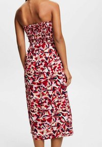 Frau, die ein trägerloses, knielanges Kleid mit rotem, pinkem und marineblauem Blumenmuster trägt, von hinten vor einem schlichten weißen Hintergrund gezeigt.