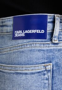 Ljust blå denimjeans med en blå etikett som står "KARL LAGERFELD JEANS." Viser synlig sömnad och detaljer i bakfickan.
