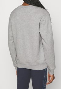 Person som bär en enkel grå långärmad sweatshirt och marinblå byxor med ett mönstrat sidostrand, vänd bort från kameran.