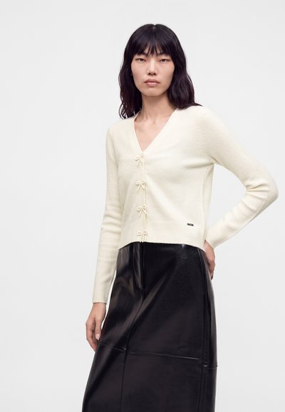 Femme aux cheveux foncés mi-longs portant un cardigan crème boutonné avec des détails nœuds et une jupe en cuir noire taille haute sur un fond uni.