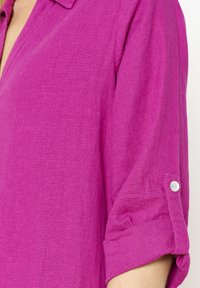 Camicia di lino fucsia con colletto e maniche a risvolto. Presenta una texture sottile e un singolo bottone bianco sulla manica.