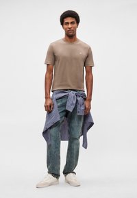 Άντρας που φοράει καφέ μπλουζάκι, μπλε καρό πουκάμισο δεμένο γύρω από τη μέση, ξεθωριασμένο τζιν και λευκά αθλητικά παπούτσια, στέκεται μπροστά σε μονόχρωμο φόντο.