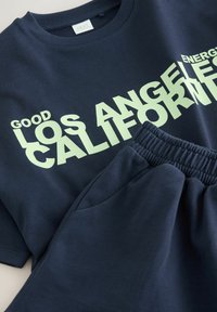 Súprava námorníckych modrých trička a teplákov. Tričko má veľký svetlozelený nápis "GOOD LOS ANGELES CALIFORNIA ENERGY." Tepláky majú elastický pás.