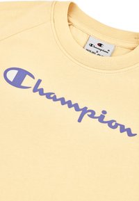 Sweatshirt en coton jaune avec un logo "Champion" violet sur le devant, avec un col rond classique et des manches courtes.