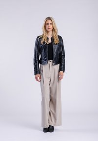 Czarna skórzana kurtka z zamkiem z przodu, zestawiona z beżowymi spodniami o szerokich nogawkach i czarnymi, spiczastymi botkami. Model stoi z neutralnym wyrazem twarzy.