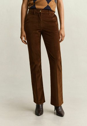 Personne portant un pantalon en velours côtelé marron taille haute associé à des bottines marron à bout pointu et talons, debout sur un fond uni.