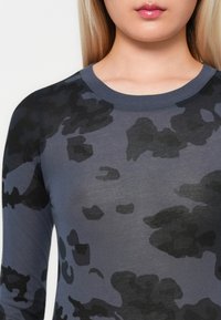 Mujer con una camisa de patrón de camuflaje negro y gris oscuro de manga larga, visible desde los hombros hasta los labios sobre un fondo simple.