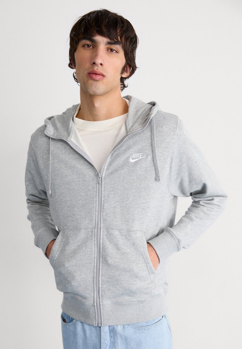 Sudadera gris con cremallera y logo de Nike en blanco, capucha con cordones, bolsillos frontales y puños acanalados, combinada con una camiseta color crema y jeans de color azul claro.