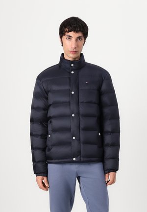 Jachetă pufă marină cu guler înalt, prevăzută cu capse și design matlasat. Modelul poartă pantaloni albaștri deschis, evidențiind un stil casual.