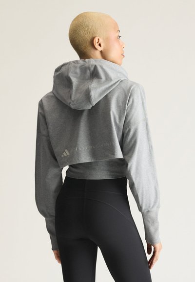 Person with short blonde hair wears gray cropped hoodie and black high-waisted leggings, standing turned slightly sideways against plain background.