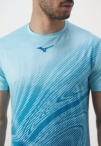 Světle modré sportovní tričko s vzorem vln, kulatým výstřihem a logem. Materiál je hladký s jemnou strukturou a vlastnostmi odvádějícími vlhkost.