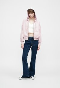 Light pink hooded sweatshirt over a white crop top, paired with dark blue flared jeans and black sneakers with white soles.