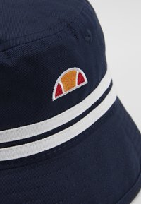 Marinblå bucket hat i bomull, med två vita ränder och en liten broderad logotyp med orangea och röda accenter.