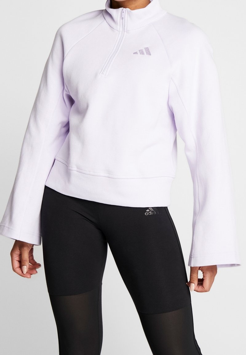 Femme portant un sweat à demi-fermeture éclair lilas à manches longues et des leggings noirs Adidas avec des panneaux en mesh sur les cuisses.