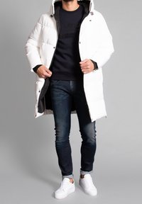 Witte pufferjas met een rits aan de voorkant, verstelbare kap en zwarte voering. Draagt over een zwart sweatshirt, gecombineerd met blauwe jeans en witte sneakers.