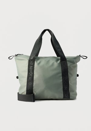 Grand sac fourre-tout en tissu vert clair avec poignées noires brandées et bandoulière ajustable, fermeture zippée sur le dessus et passants latéraux.