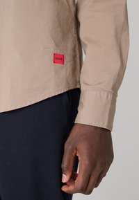 Una mano de piel oscura al lado de una camisa de manga larga beige con una etiqueta roja de "HUGO", combinada con pantalones oscuros azul marino.