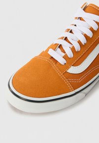 Baskets en daim orange avec bande latérale blanche, lacets blancs, et semelle en caoutchouc blanche bordée de noir, vues en angle du dessus et sur le côté.