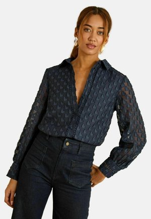 Marineblaues Hemd mit strukturiertem Herzmuster, langen Ärmeln und einem Hemdkragen mit Knöpfen. Kombiniert mit dunklen Denim-Jeans.