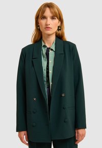 Blazer doppiopetto verde scuro con revers, caratterizzato da bottoni argentati e una vestibilità comoda, indossato sopra una camicia a righe.