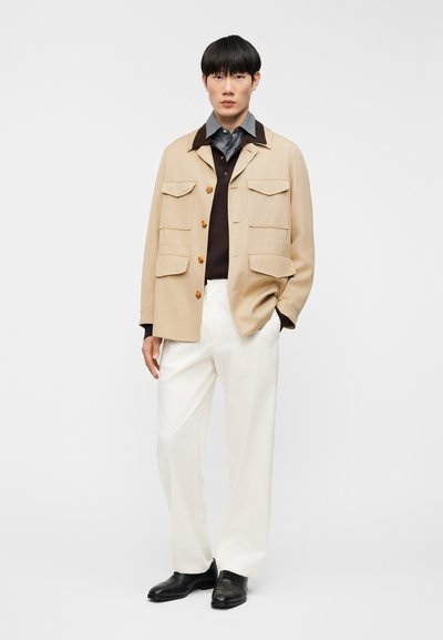 Άνδρας με μπεζ μπουφάν, σκούρο πουκάμισο, λευκό παντελόνι και μαύρα παπούτσια, που στέκεται μπροστά σε απλό λευκό φόντο.