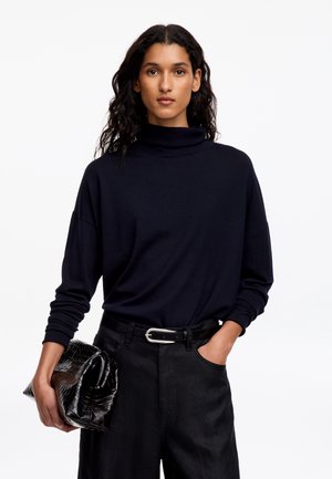 Mujer con cabello largo y oscuro, con un jersey de cuello alto negro y pantalones negros, sosteniendo un gran bolso de mano negro con textura en una mano.