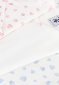 Trzy bawełniane koszule ułożone jedna na drugiej, w kolorach jasnoróżowym, białym i jasnoniebieskim, ozdobione małymi serduszkami. Delikatne wykończenia i akcenty w postaci kokardek.