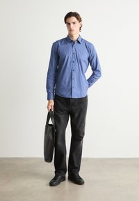 Cămașă albastră deschisă cu mâneci lungi, blugi negri și teniși negri. Ține în mână o geantă neagră. Design simplu, textură netedă.