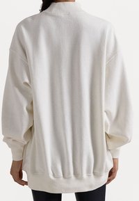 Henkilö, joka on pukeutunut suuriin yksivärisiin valkoisiin huppareihin, joissa on ribbaus hihansuissa ja helmassa, näkyy selkäpuolelta, seisten yksinkertaisen taustan edessä.