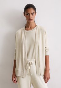 Donna con un cardigan lavorato a maglia beige sopra un top abbinato in maglia e pantaloni con coulisse, in piedi contro un muro chiaro e semplice.