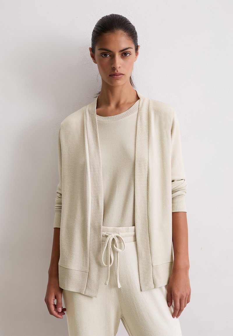Donna con un cardigan lavorato a maglia beige sopra un top abbinato in maglia e pantaloni con coulisse, in piedi contro un muro chiaro e semplice.