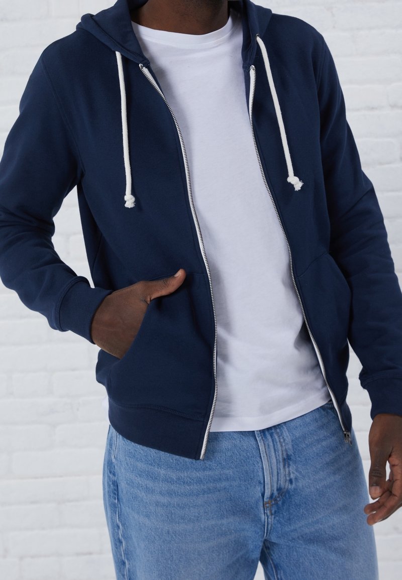 Uomo che indossa una felpa con zip blu navy sopra una t-shirt bianca e jeans blu, con una mano in tasca, davanti a uno sfondo di parete di mattoni bianchi.