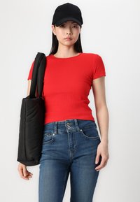 T-shirt rouge à côtes associé à un jean bleu taille haute. Casquette noire portée et portant un sac rembourré noir avec des bretelles.