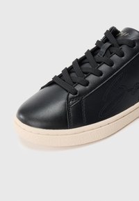 Μαύρο δερμάτινο αθλητικό παπούτσι με υφασμένο επάνω μέρος, στρογγυλή μύτη, μαύρα κορδόνια και κρεμ λαστιχένια σόλα με διακριτικές ανάγλυφες λεπτομέρειες στο πλάι.