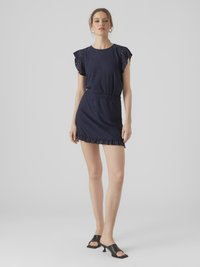 Naine, kes kannab tumesinist montloonkleiti, millel on lehvikvarrukad, ning musti avatud varbaga kontsakingi, seismas halli tausta ees.