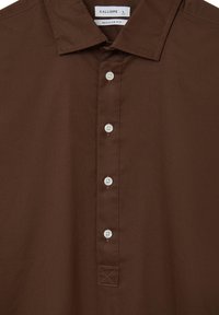 Braunes Langarmshirt mit spitzem Kragen, vier weißen Knöpfen und einem Etikett, auf dem "Calliope L Größe" und "Reguläre Passform" steht.