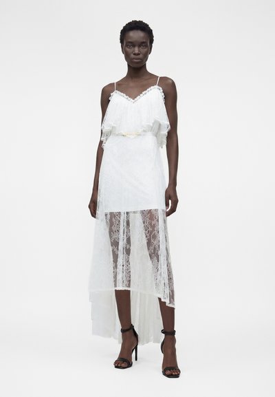 Γυναίκα που φοράει λευκό δαντελωτό φόρεμα με ψηλό μπροστινό και χαμηλό πίσω μέρος, με λεπτές τιράντες και μαύρα ψηλοτάκουνα σανδάλια, στέκεται μπροστά σε απλό φόντο.