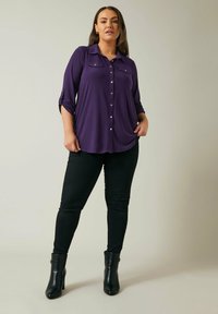 Purple button-up blouse with roll-up sleeves, two chest pockets, and a relaxed fit, paired with black skinny jeans and ankle boots.