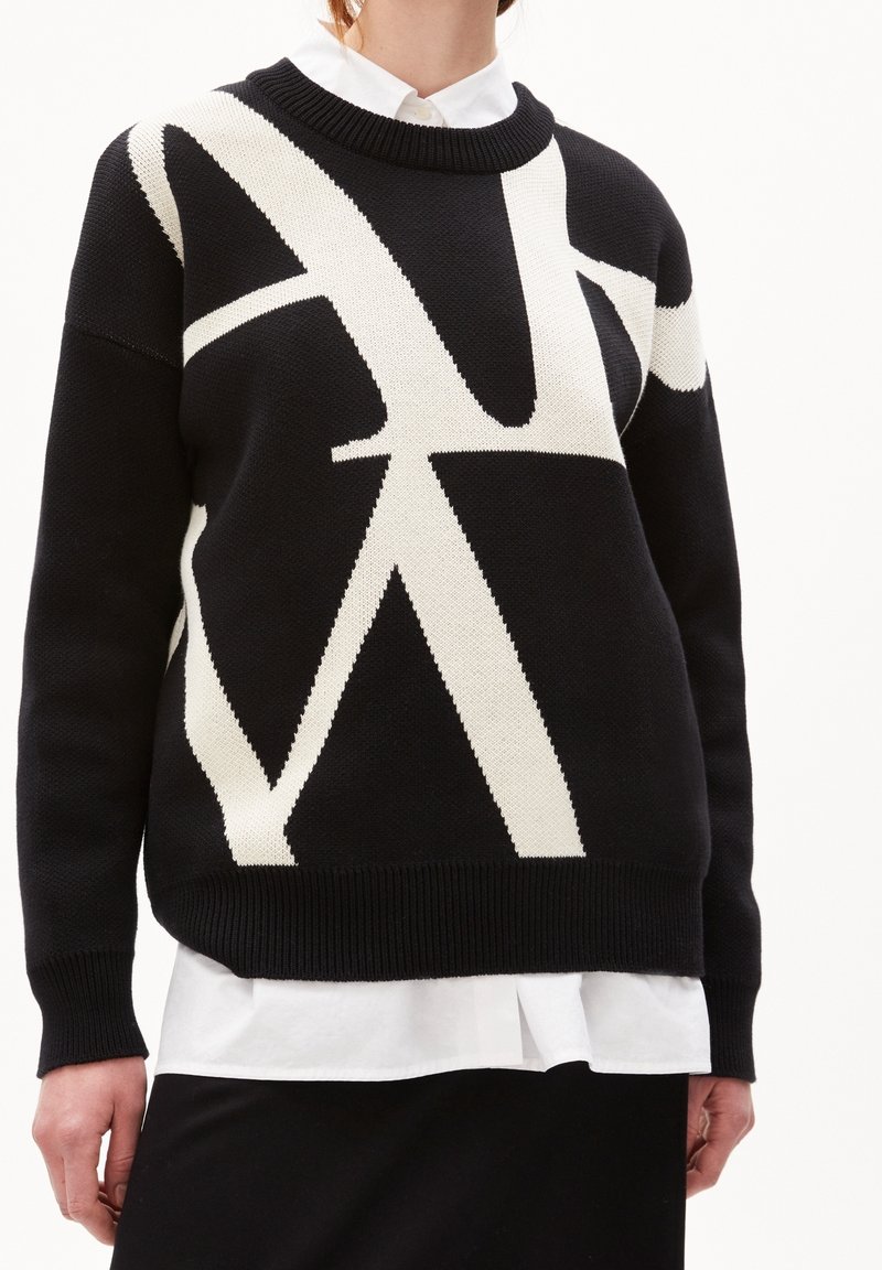 Schwarzer Strickpullover mit einem weißen abstrakten Muster, mit geripptem Kragen und Bündchen. Darunter ein weißes Hemd mit Kragen.