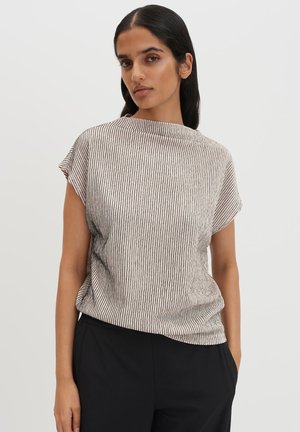 Donna che indossa una maglia beige a maniche corte con sottili righe verticali nere e pantaloni neri, in piedi davanti a uno sfondo chiaro e semplice.